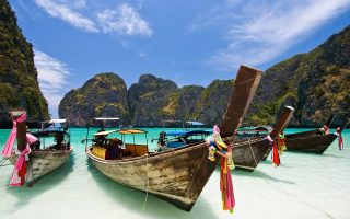 قایق‌های سنتی دم بلند که با پارچه‌های رنگارنگ تزئین شده‌اند، در آب‌های فیروزه‌ای شفاف نزدیک یک ساحل شنی لنگر انداخته‌اند. صخره های سنگ آهکی سر به فلک کشیده پوشیده از فضای سبز در پس زمینه در زیر آسمان آبی روشن با ابرهای پراکنده بالا می روند و جذابیت تجربه تور گروهی پوکت را تداعی می کنند.